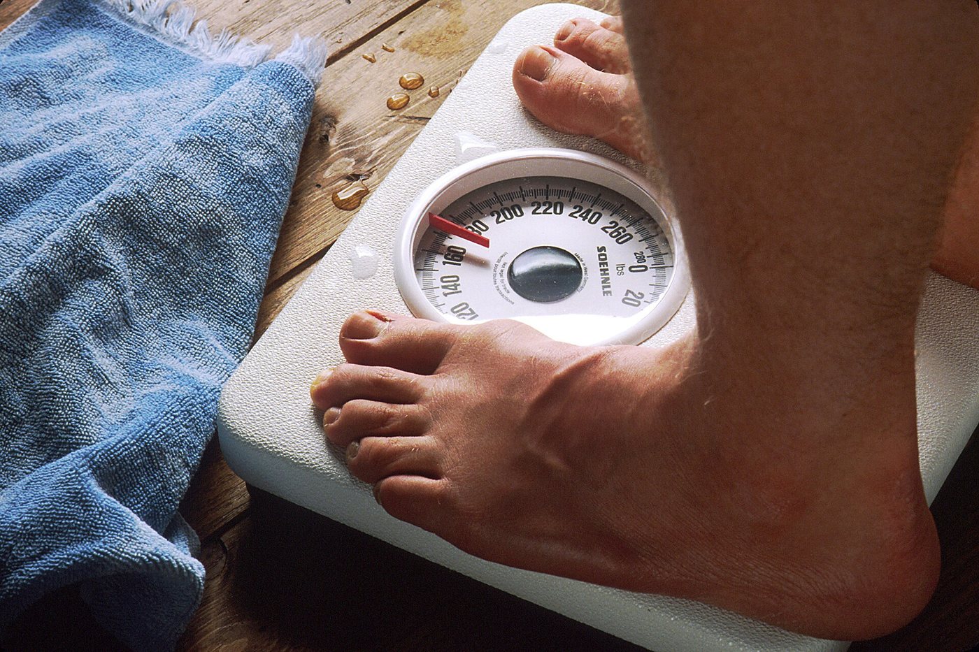Bare feet standing on an analogue bathroom scale with a round dial, beside a blue towel on a wooden floor.