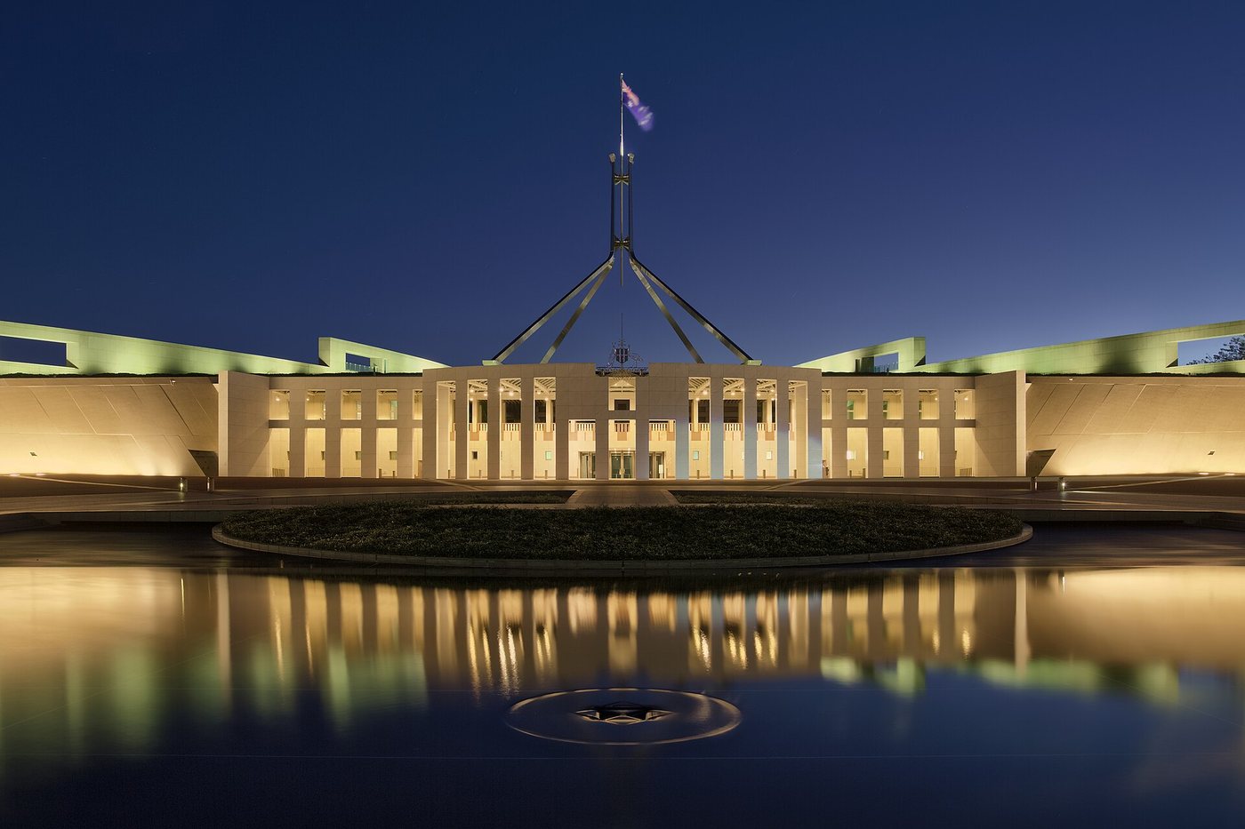 Parliament House in Canberra, Australia, at dusk, with its tall flagpole and lit facade reflected in the forecourt water feature.