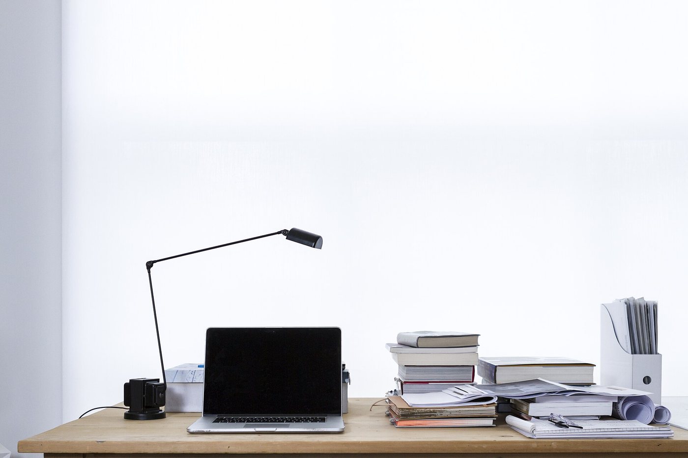 An open laptop on a light wooden desk beside a desk lamp and stacks of books, set against a bright white wall.