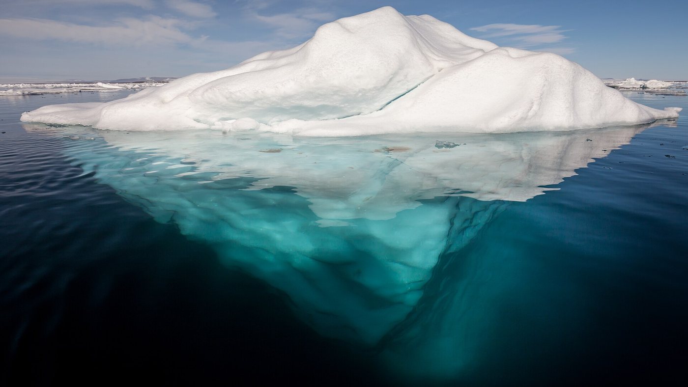 An iceberg floating in calm, clear Arctic water, with its much larger submerged underside visible below the surface.