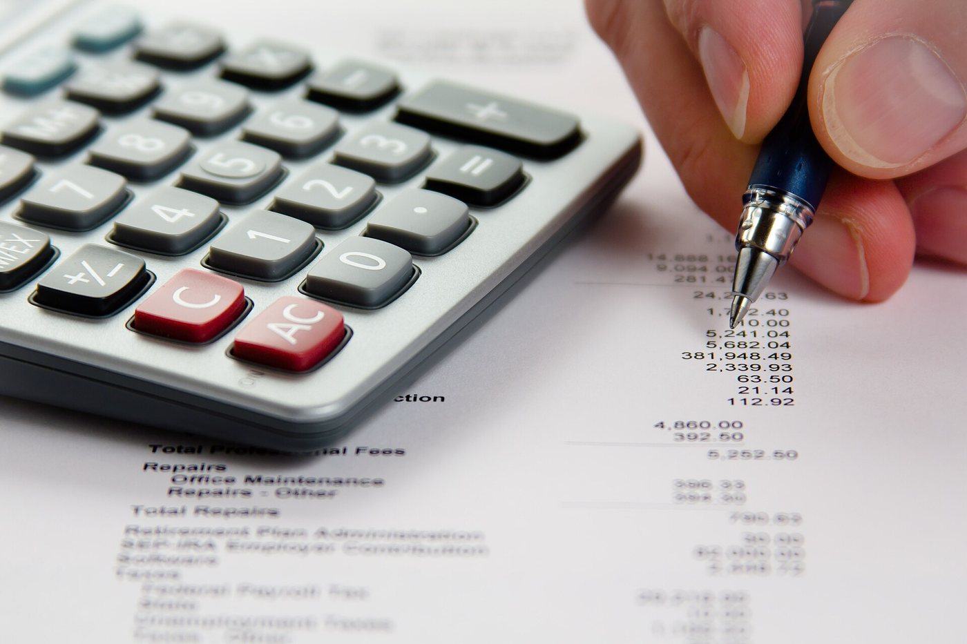 Close-up of a hand with a pen checking figures on a printed financial statement next to a desktop calculator.