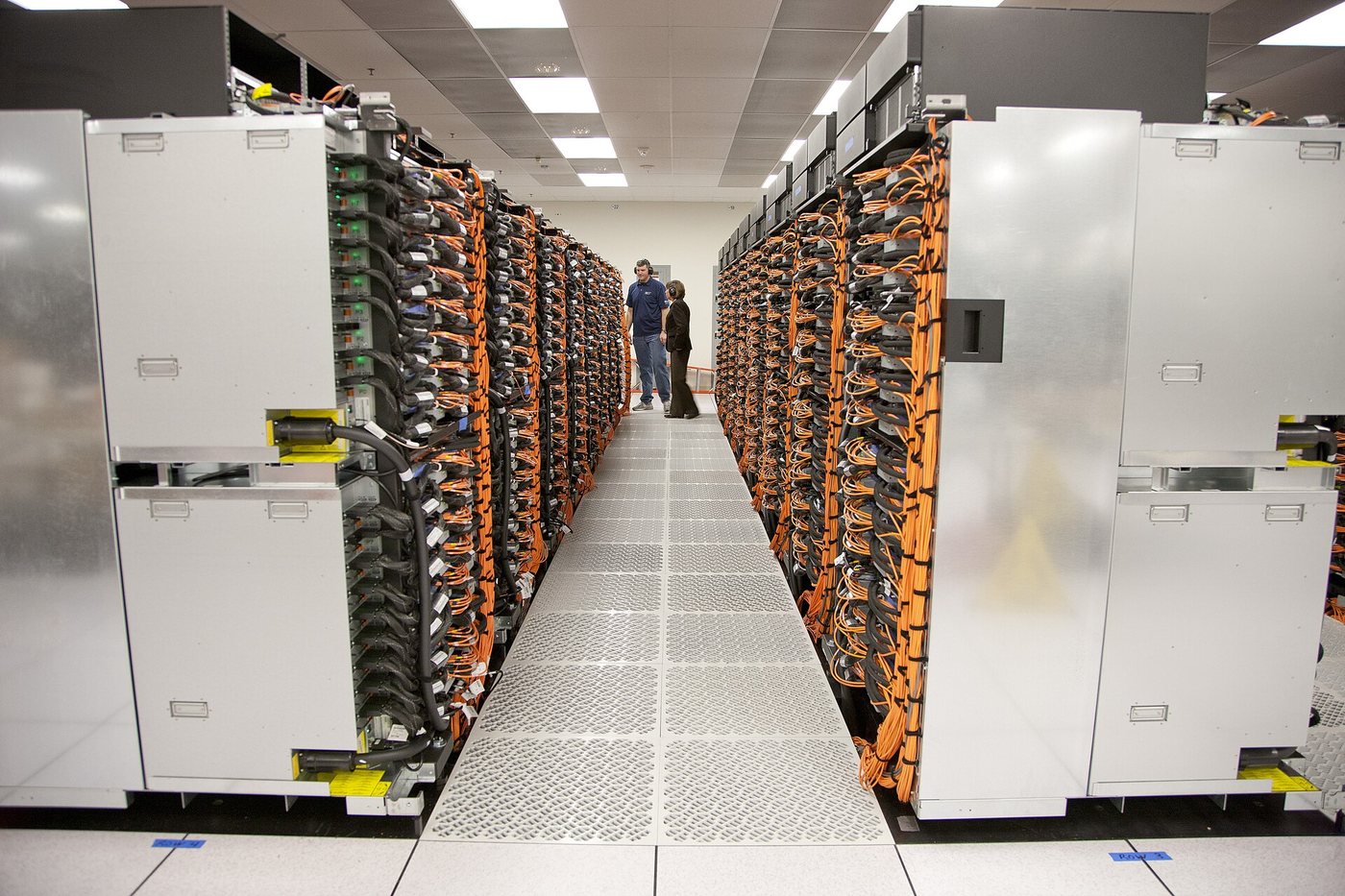 A long aisle between rows of racks of the Sequoia supercomputer at Lawrence Livermore National Laboratory, with orange cabling and two small figures at the far end.